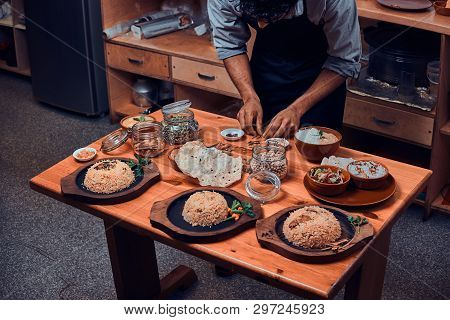 Cook Prepares Spices From Jar For His Indian Dishes. There Are Few Wooden Plates With Rice, Jars Wit