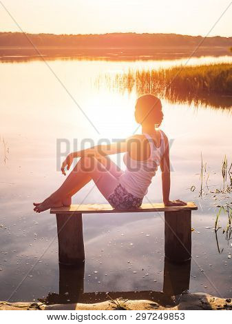 Pensive Girl Sits On A Bench By The River And Enjoys A Beautiful Sunset. Girl Sitting Near Water Out