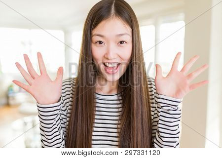 Beautiful Asian woman wearing stripes sweater over living room background celebrating crazy and amazed for success with arms raised and open eyes screaming excited. Winner concept