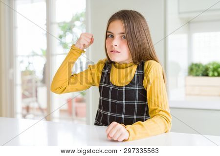 Young beautiful blonde kid girl wearing casual yellow sweater at home angry and mad raising fist frustrated and furious while shouting with anger. Rage and aggressive concept.