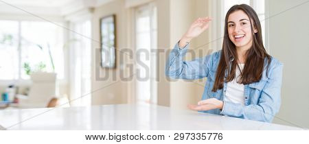 Wide angle picture of beautiful young woman sitting on white table at home gesturing with hands showing big and large size sign, measure symbol. Smiling looking at the camera. Measuring concept.