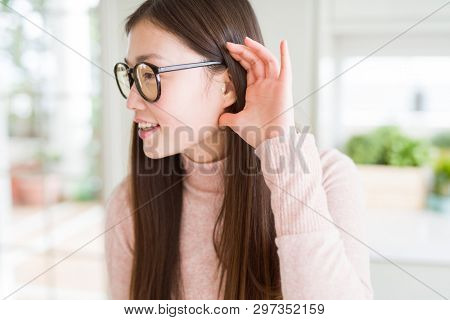 Beautiful Asian woman wearing glasses smiling with hand over ear listening an hearing to rumor or gossip. Deafness concept.