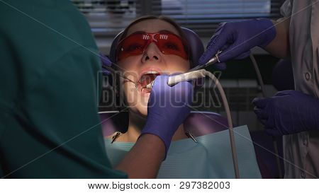 Dentist Is Taking Care About Patients Tooth Using Electric Dental Brush. Nurse With Saliva Ejector N