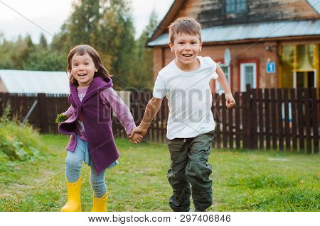 Brother And Sister Run By The Hand In The Background Village House. Holidays In The Village With My 
