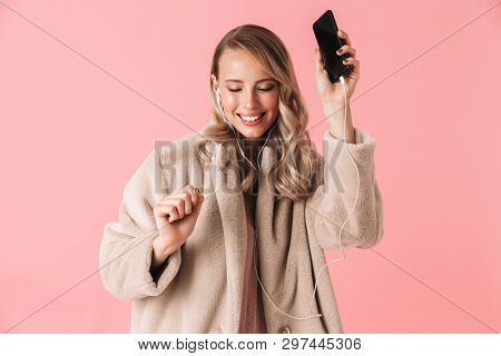 Pleased blonde woman wearing in fur coat listening music and dancing with closed eyes over pink background