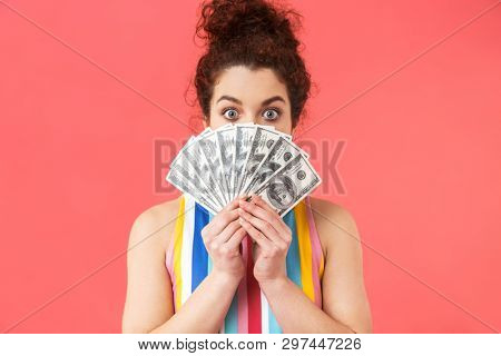 Playful pretty redhead woman wearing in dress hiding behind money and looking at the camera over pink ackground