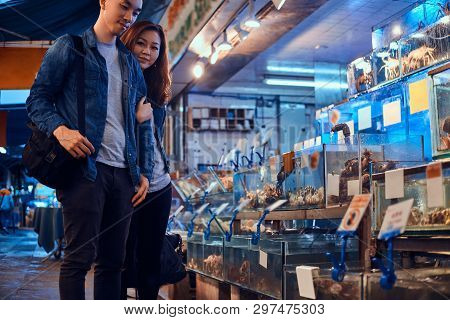 Young Asian Couple Looking For Something Special At Traditional Chainese Market. They Are Pensive. A