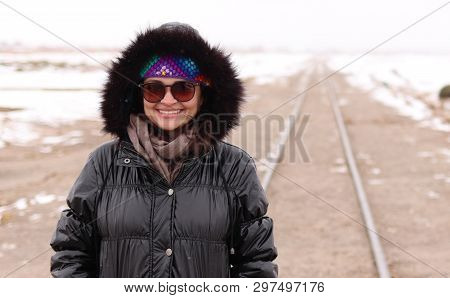 Smiling White Girl Wearing Sunglass, Black Coat, Brown Scarf, Colorful Band In Front Of A Train Trai