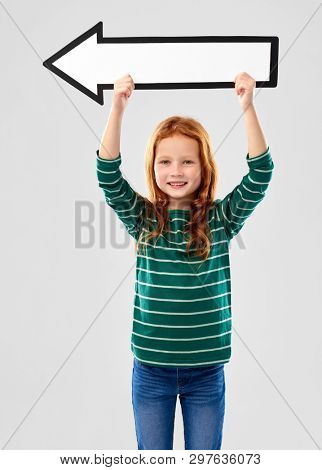 childhood, fashion and people concept - smiling red haired girl in green striped shirt holding big white leftwards thick arrow over grey background