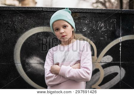 A Serious Child Girl Folded Her Arms Across Her Chest Against The Background Of A City Wall With Gra