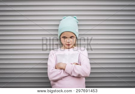 A Serious Child Girl Folded Her Arms Across Her Chest Against The Background Of A City Wall With Gra