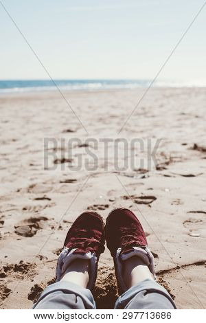 Feet Of Young Woman In Sport Footwear And Jeans Relaxing After Hiking On Sand Beach. Pov. Adventure 