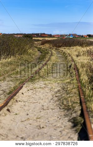 A Rusty Railroad Track On A Sandy And Grassy Ground Leading To A Small Village In Germany.
