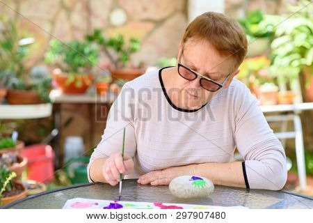 Happy Authentic Senior Woman Sitting Outdoors At Table Painting Pictures On Stone.