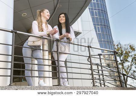 Two Beautiful Women Are Talking At A High-rise Glass Building, Leaning On The Railing, View From Bel