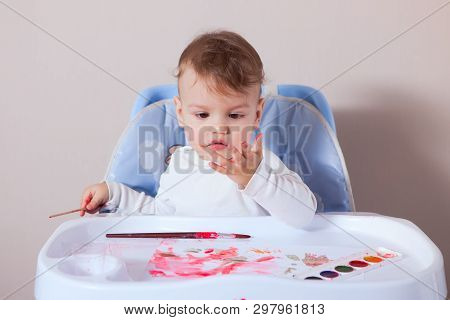 A One-year-old Child, Smeared In Paints, Sits At A Children's Table And Looks At The Dirtied Hands.