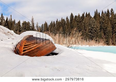 Old Wooden Destroyed Flooded Fishing Boat Lie In Snow On Shore Of Ice-covered Frozen River In Winter