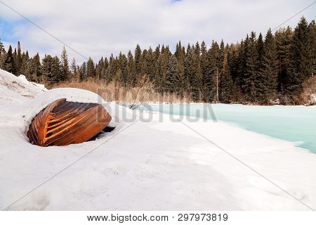 Old Wooden Destroyed Flooded Fishing Boat Lie In Snow On Shore Of Ice-covered Frozen River In Winter