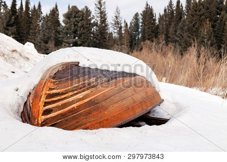 Old Wooden Destroyed Flooded Fishing Boat Lie In Snow On Shore Of Ice-covered Frozen River In Winter