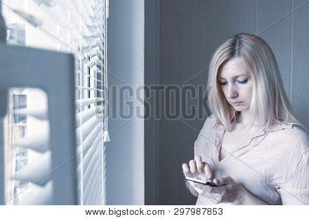 Sad Apathetic Lonely Woman Standing Near The Window And Texting Messages In Smartphone At Home Or Ho