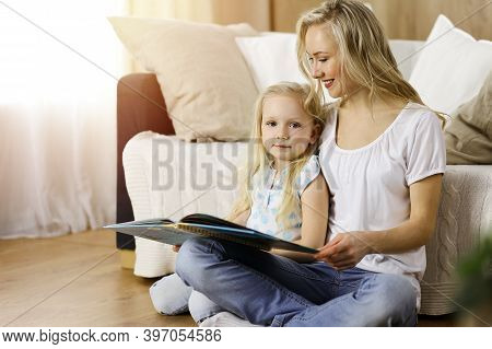Happy Family. Blonde Young Mother Reading A Book To Her Cute Daughter While Sitting At Wooden Floor 