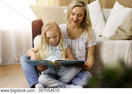 Happy Family. Blonde Young Mother Reading A Book To Her Cute Daughter While Sitting At Wooden Floor 