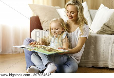 Happy Family. Blonde Young Mother Reading A Book To Her Cute Daughter While Sitting At Wooden Floor 
