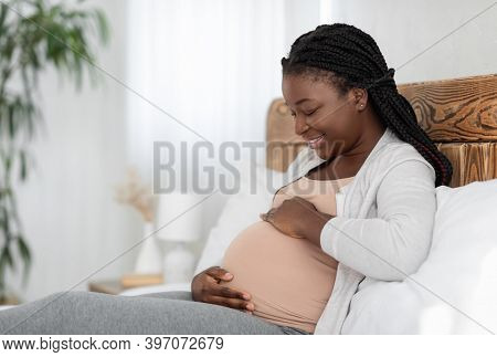 Cheerful Black Pregnant Woman Relaxing On Bed At Home, Touching Belly And Smiling, Looking At Her Bi