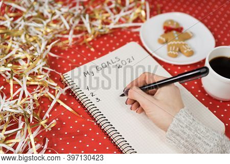 Female Hands Writing My Goals 2021 In A Notebook. Gingerbread Man, Coffee, Red Background And Tinsel