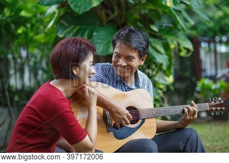 Middle Aged  Couple Playing Guitar While Relax Sitting On Bench In Backyard.
