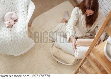 Woman In Robe Is Sitting Near Wooden Shelving, Organizing Closet By Sorting And Folding Bath Towels.