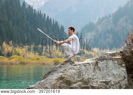 A Barefoot Man Sits On A Stone And Holds A Sword In His Hands. Preparing For The Battle. Man Health 