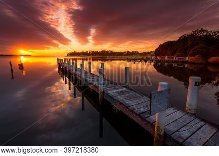 Old Weathered Timber Jetty Pointing Towards The Sunrise With Pretty Reflections In The Water. Mallac