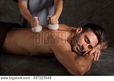 A Good-looking Man Getting A Back Massage Lying Down At Spa, Pouches Filled With Dry Herbs Are Brush