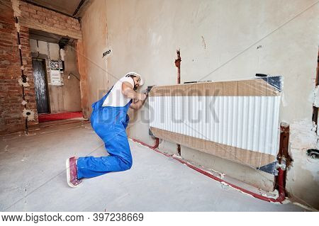 Plumber In Work Overalls Using Wrench While Installing Heating Radiator In Room. Man In Helmet Insta