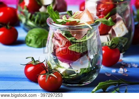 Salad With Arugula, Cheese, Cherry Tomatoes And Prosciutto In Glass Jars, Pink Blue Background, Sele
