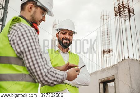 Low Angle Of Smiling Multiracial Male Coworkers In Hardhats And Waistcoats Using App On Mobile Phone