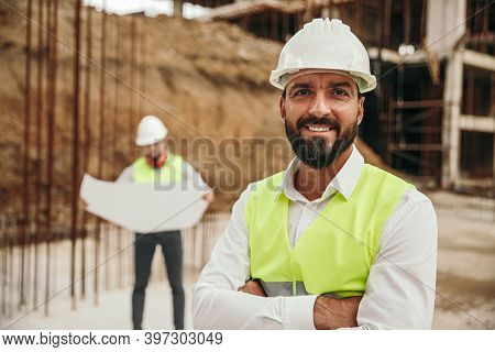Positive Confident Ethnic Bearded Male Construction Worker In Hardhat And Waistcoat Looking At Camer