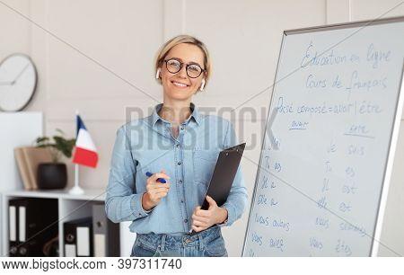 Friendly Young Female Tutor Standing Near Blackboard With French Grammar Rules, Giving Web Lesson Fr
