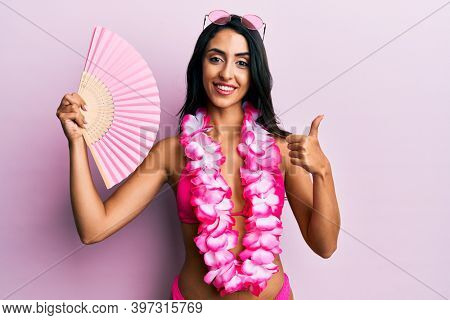 Beautiful hispanic woman wearing bikini holding handfan smiling happy and positive, thumb up doing excellent and approval sign 