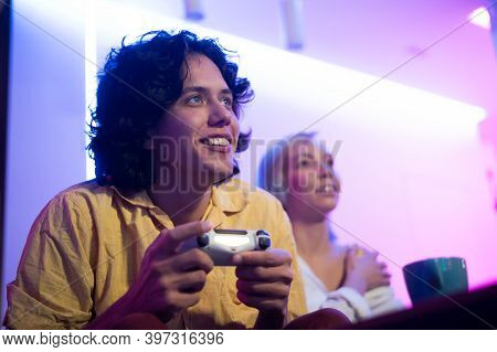 Young Curly Man And His Girlfriend Playing Gaming Console Using Joystick At Home Lit With Neon Pink 