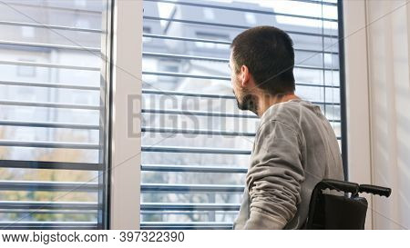 Man On Wheelchair In Living Room Looking Through Window. Handicapped Invalid Paralysed Person, Depre