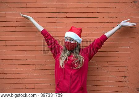 Blond Girl In Christmas Costume And Red Protective Mask Stands With Hands Raised Up. Welcome And Joy