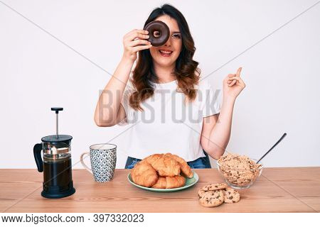 Young beautiful brunette woman eating breakfast holding cholate donut smiling happy pointing with hand and finger to the side 