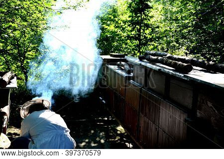 The Beekeeper Kneels In A White Jumpsuit With A Hood And A Hat To The Smoker Who Must Light To Burn 