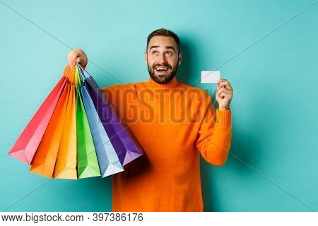 Happy Aduly Man Showing Credit Card And Shopping Bags, Standing Against Turquoise Background