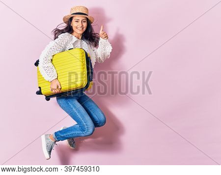 Young beautiful woman on vacation wearing summer clothes and hat smiling happy. Jumping with smile on face holding cabin bag over isolated background