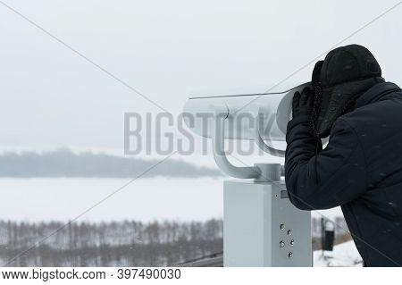 A Man In Warm Clothes Looks Through A Binoscope At The Winter Landscape. Copy Space.