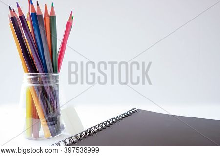 Colored Pencils In A Transparent Glass And A Black Notebook On An Isolated White Background