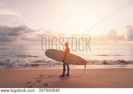 Man Hold Surfboard Standing At Tropical Sunset Beach Background. Summer Vacation And Sport Adventrue